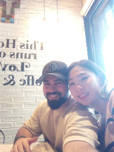 A selfie of a man and a woman in an indoor environment with white brick walls. The man is wearing a New York Yankees baseball cap and a tan shirt. He has a dark beard and a bracelet on his left wrist. The woman is wearing a pink top and has fair skin and short blonde hair. Behind them is a wall with large black block lettering. There is a window to the right, letting in a lot of light. The specific location within Valencia, Venezuela cannot be determined based on the visual information provided.
