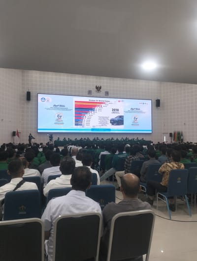 The image shows a large indoor gathering in a spacious hall, likely an auditorium or conference center. A prominent, wide screen displays a presentation titled "Global EV Brand Sales" with bar charts showing sales figures for various car brands and the year 2018. The screen also features logos and text in what appears to be Indonesian. The audience, composed of many individuals, is seated in rows of chairs facing the screen. The attendees are dressed in a mix of casual and formal attire, with some wearing uniforms. Some audience members are wearing green shirts, while others are in white shirts, and some are in batik. A few individuals are standing in the front, near a podium where a speaker is present. The hall has a high ceiling with lighting fixtures. The overall atmosphere suggests a formal event, possibly a conference, seminar, or university assembly. The location is identified as Reuleut Timu, Indonesia, based on the text displayed on the screen, which includes "SENAT UNIVERSITAS MALIKUSSALEH" and "REULEUT".