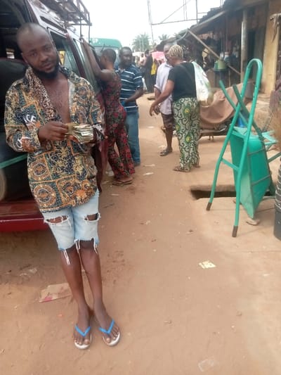 The image portrays an active street scene in Nnobi, Nigeria, characterized by reddish-brown unpaved ground. In the immediate foreground, a dark-skinned adult male with a shaved head and beard stands, leaning against the side of a dark-colored vehicle, possibly a minivan, which features an overhead luggage rack. He wears an unbuttoned, multi-patterned long-sleeved shirt, light-colored distressed denim shorts with rips, and white flip-flops with blue straps. He holds a substantial stack of banknotes in his hands, facing the viewer.
To the right of the foreground subject, a group of individuals is gathered. This group includes a female with short hair, wearing a multi-colored floral jumpsuit, who appears to be interacting with the open door of the vehicle. Directly behind her stands a male in a striped polo shirt and blue jeans. Further to the right, another male in a light-colored short-sleeved shirt is observed conversing with a female dressed in a black t-shirt and green patterned trousers; she carries a large white sack on her right shoulder.
The midground features a prominent green metal object, resembling a wheelbarrow frame or a trolley, positioned near a square-shaped opening in the ground. A broom with dark bristles is partially visible adjacent to the green object. The background consists of several single-story structures, consistent with market stalls, topped with corrugated metal roofing. Multiple indistinct figures are visible further down the street, suggesting a busy public area. The overall lighting indicates an overcast sky.