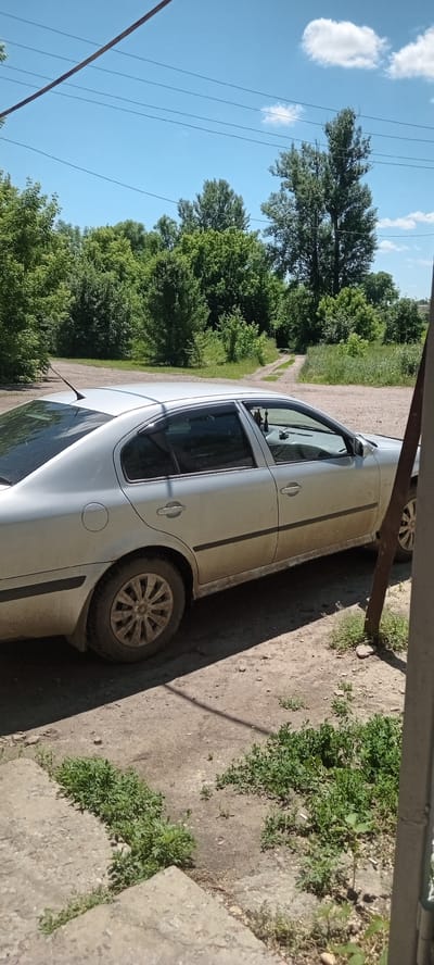 A silver sedan is parked on a dirt road surrounded by trees and a blue sky with a few clouds. The car appears to be in a rural or suburban area, possibly Koviagy, Ukraine, given the natural scenery. The car has some dirt on its lower panels and wheels, suggesting it has been driven on unpaved surfaces. The scene is illuminated by natural daylight, casting shadows on the ground. There are power lines visible in the upper portion of the image, stretched across the sky. In the foreground, there are concrete steps and patches of green vegetation. A metal pole is partially visible on the right side of the image.