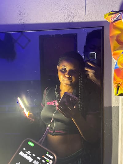 The scene, an indoor mirror reflection captured in Gbongan, Nigeria, is illuminated by multiple artificial light sources, primarily from smartphones. A dark-skinned female, reflected from the chest up, stands centrally. She wears a black, short-sleeved crop top with pink stitching and a zipper, revealing her midriff. She has braided hair, a nose piercing, and holds a smartphone with a pink case in her right hand. A second dark-skinned individual, likely male, is partially reflected behind her, holding a third smartphone with a visible multi-lens camera system, indicating he is taking the photograph. A second smartphone, in the lower-left foreground, emits a bright light and partially displays a screen showing "Activity All." The mirrored background reveals a dark blue wall with a faint, reflective geometric outline on the left and a darker rectangular shape on the right. To the immediate right of the mirror, a colorful bag of "Fun Snax" is visible against a light-colored wall, beneath a speckled surface above the mirror.