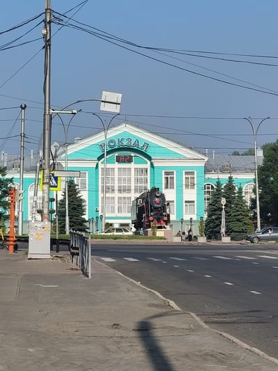 The image shows the Kemerovo railway station in Kemerovo, Russia, under a clear blue sky. The station building is painted in a distinctive turquoise and white color scheme. A vintage steam locomotive, painted black with red accents, is displayed in front of the station as a monument. A large digital clock displaying "82" is visible on the facade of the station. On the sidewalk, there are several people, and a traffic light is visible to the left. The street in front of the station has a crosswalk and a few cars. The overall scene is that of a public space, likely a transportation hub, on a bright, sunny day.