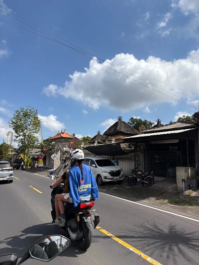 In Tampaksiring, Indonesia, two individuals ride a scooter along a paved road under a bright blue sky with scattered clouds. The person in the passenger seat, wearing a blue sweatshirt with the word "OUTSIDE" in white lettering and denim shorts, has their hair in a braid. They are also wearing a white helmet. The driver, partially obscured, wears a dark shirt and is also on the scooter. Several vehicles, including a white car and a motorcycle, are parked on the side of the road. Buildings with traditional Balinese architecture, characterized by their peaked roofs and ornate facades, line the street. The scene captures a moment of daily life in a tropical urban environment, with lush green trees visible in the background.