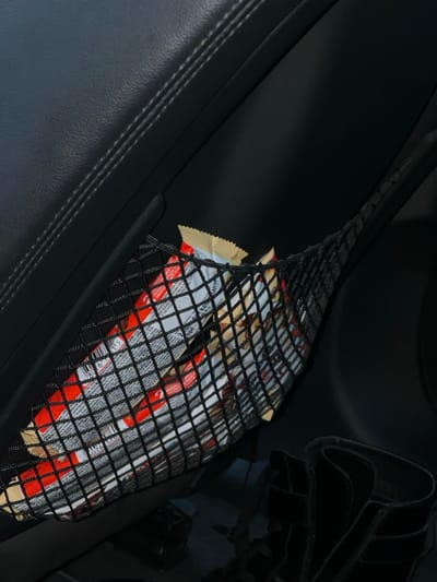 The scene captures the interior of a vehicle, specifically a dark, textured panel—likely a door panel or seat-back—exhibiting parallel stitching. A black mesh storage net is affixed to this panel. The net is filled with numerous individually wrapped snack bars, predominantly featuring red and white/silver packaging. Several wrappers bear discernible "Kinder" branding, barcodes, and text, suggesting products such as Kinder Bueno. Below the net, dark, amorphous objects are partially visible on the vehicle's lower interior. No persons are present. The precise location within Stara Pazova Municipality, Serbia, cannot be ascertained from the visual evidence provided.