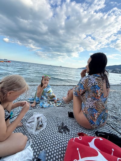 The image depicts three individuals at a beach, with the sea and a cloudy sky in the background. The scene appears to be set in Yalta, Ukraine.
In the foreground, a young girl with blonde hair is sitting and looking down, seemingly engaged with something in her hands. To her left and slightly behind her, another child, possibly a girl, is also sitting, wrapped in a blue and white towel. She is wearing green goggles and is looking up and to the left.
In the background, an adult with long dark hair is crouching and looking down. She is wearing a colorful, patterned dress with a leopard-like print. The individuals are sitting on a patterned blanket spread out on a pebble beach. A red towel with white anchor patterns is visible on the right side of the image. A clear plastic bag filled with what appears to be sunflower seeds is also present on the blanket.
The sky is filled with a mix of blue sky and dense clouds, suggesting it might be a partly cloudy day. The sea is calm, with gentle waves. The overall impression is a relaxed, casual outing at the beach. No visible text is present in the image.