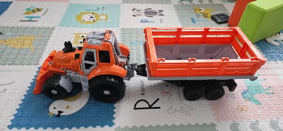 An orange toy tractor with a grey and orange trailer sits on a multi-colored mat. The mat features various animals and letters. The tractor has large black wheels and a detailed cab. The trailer is open at the top and has orange railing. A black calculator and green and orange blocks are visible in the background, suggesting an indoor play area. The scene is a static, close-up shot. The image is taken from a high angle.
