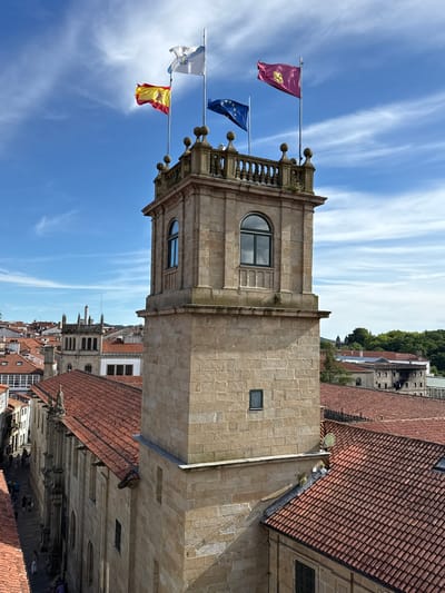 The image shows a view from above in Santiago De Compostela, Spain, focusing on a stone tower adorned with several flags. The flags visible are the Spanish flag, the European Union flag, a white flag with a star and possibly a symbol, and a maroon flag with a yellow emblem. The tower is constructed from large, light-colored stones and features arched windows with dark panes and decorative stonework. Below the tower, red-tiled rooftops dominate the scene, indicative of traditional Spanish architecture. On the left side of the image, a narrow street is visible with several people walking along it. The sky is blue with scattered white clouds. The overall scene captures a historic urban environment.
