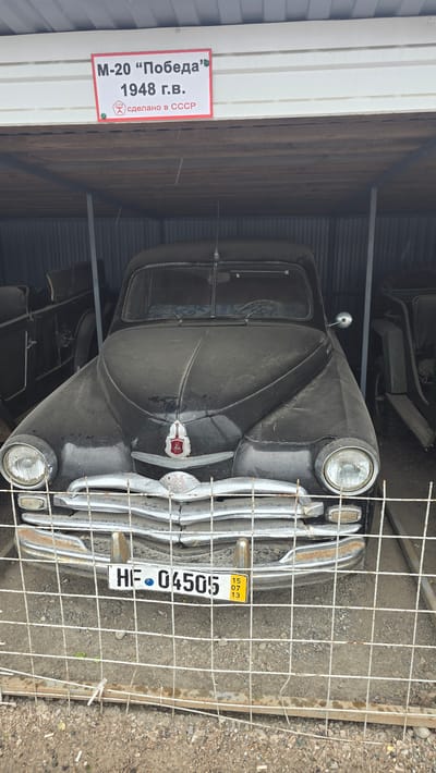 The image shows a dusty, black Soviet-era car, identified as an M-20 "Pobeda" from 1948, parked under a metal roofed shelter. The car has a chrome grille, round headlights, and a red emblem on its hood. A license plate reading "HF 04505" is visible on the front. A small yellow sticker with "15 07 13" is affixed to the license plate. The car is behind a wire fence. Other vehicles, appearing to be vintage military jeeps or trucks, are partially visible in the background to the left and right of the M-20. The ground is covered in gravel and dirt. A sign above the car reads "M-20 'Победа' 1948 г.в. сделано в СССР" which translates to "M-20 'Victory' 1948 year of manufacture. Made in the USSR". The location is stated to be in the city of Bol'shoi Mashlyak, Russia.