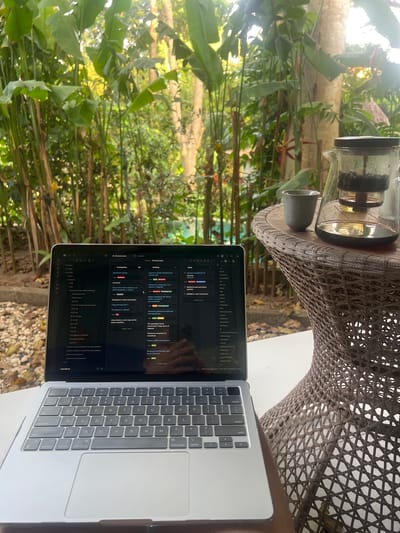 The image shows a laptop on a table in an outdoor setting, possibly a garden or patio, in Kerobokan, Indonesia. The laptop screen displays what appears to be a project management software interface in Russian. A coffee brewing apparatus and a small cup sit on the same table. The environment includes lush green foliage and plants. No people are visible.