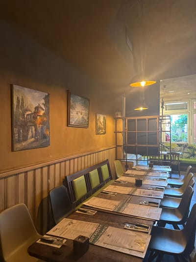 This is a dimly lit interior shot of a restaurant, likely in Sankt Wendel, Germany. The perspective is from a low angle, looking upwards and across a long table laden with menus and place settings. Several chairs, some with bright green upholstery, are visible along the table. In the background, two industrial-style pendant lights hang from the ceiling, casting a warm glow.

Further back, a large, industrial-style metal shelving unit is prominent. Beyond this, through a large window or doorway, a glimpse of greenery and trees suggests an outdoor area or garden. The walls are painted a warm, earthy tone. Several framed landscape paintings hang on the wall, depicting pastoral scenes.

The table is set for dining, with menus prominently displayed. The visible text on the menus includes "HASENKARTE," "SALATE," "PIZZEN," and "KLEINIGKEITEN," indicating a German restaurant menu. The scene appears to be set up for customers, but no people are actively engaged in dining or any activity. The lighting suggests it might be evening or a generally dimly lit indoor space. The overall atmosphere is one of readiness for service, with a rustic yet industrial aesthetic.