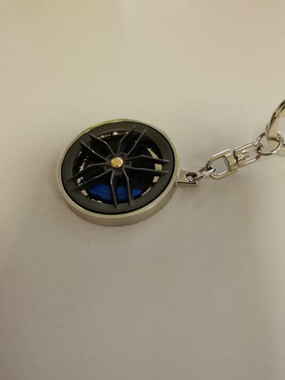 A close-up, high-angle shot shows a keychain with a car wheel design on a light-colored, textured surface. The keychain features a circular charm with a detailed, dark gray replica of a car wheel with five spokes and a gold center cap. Behind the spokes, a bright blue element, possibly representing a brake caliper, is visible. The charm is attached to a silver chain with a lobster clasp. The lighting is soft, casting subtle shadows and highlighting the metallic sheen of the keychain. The surface beneath appears to be a smooth, matte material.