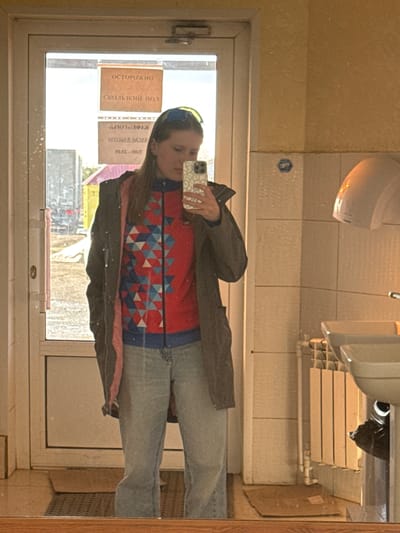 A person is taking a selfie in a bathroom mirror. The person is wearing a red, blue, and white geometric patterned sweater, jeans, and a dark jacket. The bathroom has white tiled walls and a sink. A radiator is visible above the sink. The mirror reflects a view of the outside, including a street with vehicles and signage in Russian. One sign reads "ОСТОРОЖНО СКОЛЬЗКИЙ ПОЛ" which translates to "CAUTION SLIPPERY FLOOR." Based on the sign and signage from a shop in the background, the location is the city of Lunezhki, Russia.