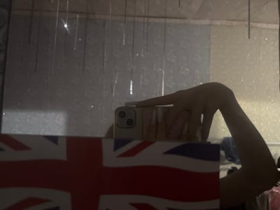 A person's arm and hand are visible in the foreground, holding a smartphone in front of a mirror. The phone case has a pink and white checkered pattern. The arm is angled upwards and to the right. In front of the hand and phone, a portion of the Union Jack flag is visible, obscuring the lower part of the scene. The background is a reflection in the mirror, showing a patterned wallpaper and vertical lines, possibly from blinds or light fixtures. The overall lighting is dim with some reflections. This scene is not in Velykyi Sukhodil, Ukraine.