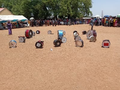 The image shows a sandy field with a collection of drums, all lying on their sides. Most of the drums are in the foreground, and several are in the middle ground. There are also people in the background, standing in a crowd, and the image appears to be taken in an outdoor setting. The coordinates (latitude: 10.116, longitude: 13.046) indicate a location in Zum, Nigeria. The area surrounding the drums is covered in sand, with little vegetation. The people are positioned behind a white tent with a blue awning. They appear to be observing the drums, which are likely awaiting a performance. The people are dressed in a variety of clothes, ranging from casual to formal. Some people are wearing hats, while others are not.
