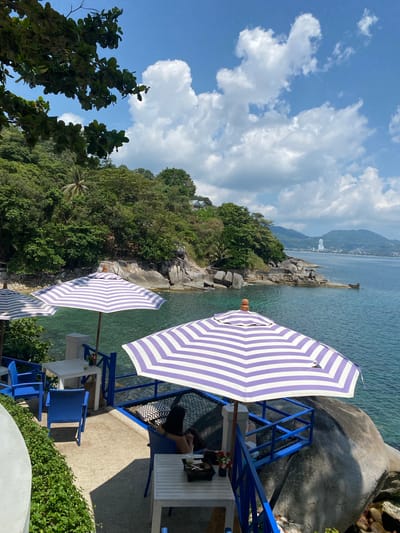 The image shows a scenic view, likely taken at a coastal resort or beachside establishment in Kamala, Thailand. In the foreground, there are several blue-and-white striped beach umbrellas and blue outdoor furniture set up on a patio or terrace. One person is visible, seated near a white table.
The midground features the turquoise ocean meeting a rocky shoreline with lush, green vegetation covering the hillside. In the background, there is a distant view of the coastline with buildings and possibly other resorts or structures along the shore. The sky is blue with scattered white clouds, indicating a sunny day.