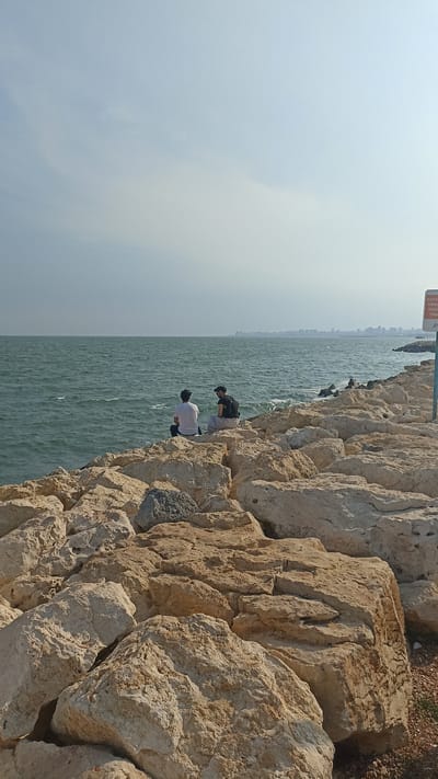 Two individuals are seated on a rocky shoreline, facing a body of water. The person on the left is wearing a white t-shirt and dark pants, while the person on the right is wearing a black t-shirt and a dark cap. They are situated on a breakwater composed of large, light-colored rocks. The sea is choppy with visible waves, and a hazy skyline of a city is visible in the distance. The sky is mostly clear with some faint clouds. A sign is partially visible on a pole to the right of the image, indicating a warning or information. This scene appears to be located on the coast of Yenişehir, Turkey, a region known for its coastal areas and urban developments along the water.