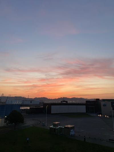 The image presents a twilight cityscape. The sky exhibits a gradient of color from a faint blue to orange. The horizon contains indistinct structures, including buildings and cranes, suggestive of a harbor or industrial zone. Adjacent to this scene lies a paved area, alongside modern architectural structures, including buildings and possibly park facilities. Adjacent to the built environment, there is green space, possibly a park or grassy area, where a person stands. The scene is in Gijón/Xixón, Spain.