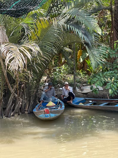 Two men are seated in two boats in Tân Thạch, Vietnam. One boat, light blue with red and yellow accents, is occupied by a man wearing a gray shirt, blue jeans, and a dark hat, appearing to use a cell phone. The other boat is dark blue and holds a man with bare feet, wearing a white shirt, dark pants, and a white hat. The boats are docked alongside lush vegetation, including large palm leaves. The water is murky.