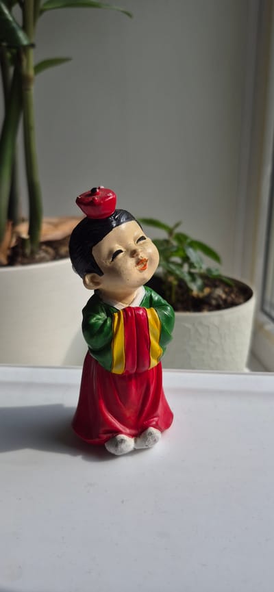 A small, ceramic figurine of a child in traditional Korean clothing is positioned on a white, flat surface. The figurine has black hair, a painted face with eyes closed and mouth slightly open, and wears a red hat with a black dot on top. Its garment is primarily red, with green sleeves and yellow and red stripes on the chest area. The figurine's feet are visible, appearing to be white. The background consists of a blurry indoor environment with a large green plant in a white pot to the left and another green plant in a white pot to the right. A window is visible in the background, casting shadows from the figurine onto the surface it stands on. The lighting suggests it is daytime, with sunlight streaming in. The image does not provide any information to suggest its location in Ussurijsk, Russia.