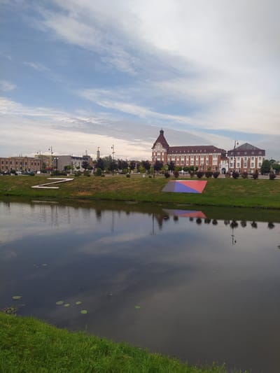 The image captures a waterfront scene in Gusev, Russia, featuring a river or canal in the foreground, with buildings and a grassy embankment lining the opposite side. The water reflects the sky, clouds, and surrounding structures. On the grassy embankment, a large, stylized letter "Z" is formed, composed of white elements. Further along the embankment, a large, colorful geometric art installation is visible, with sections of red, blue, and purple. Beyond the art installation and the embankment, a row of buildings is present. The most prominent building is a multi-story, brick-colored structure with a distinctive turret and numerous windows, suggesting it might be a historical or institutional building. To its right, another building with white facades and dark roofing is visible. The sky above is partly cloudy, with a mix of blue sky and white clouds. The overall atmosphere is calm and somewhat overcast, indicative of a day with variable weather.