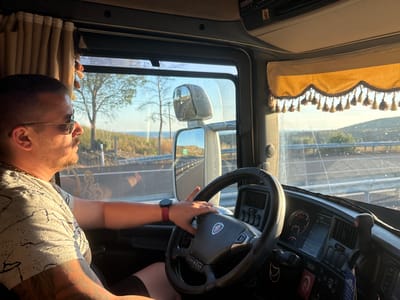 The image shows a man driving a truck. He is wearing sunglasses and a t-shirt. The truck's interior is visible, including the steering wheel, dashboard, and a decorative curtain. The man's right arm is on the steering wheel, and his left arm is resting on the door. The view outside the window shows a road, trees, and hills under a clear sky. The scene appears to be set in Avra, Greece, based on the provided context.