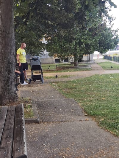 The image shows a park scene in Troyan, Bulgaria, with a man in a yellow t-shirt and black shorts standing next to a stroller. A person, likely a woman based on posture, is seated on a bench to the left. The ground is paved with concrete and there is a grassy area with several pigeons scattered around. Mature trees with dense foliage are present, and in the background, a building with windows and a fence can be seen.