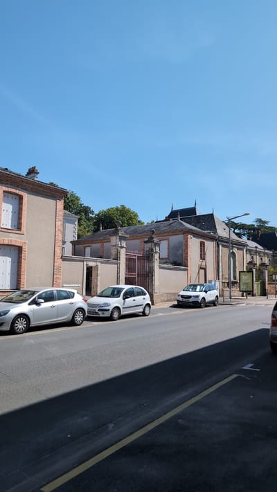 The image shows a street in Cholet, France, with several cars parked along the side of the road. In the background, there are buildings with distinctive architectural features, including a large gate with ornate ironwork. The sky is clear and blue, indicating a sunny day. The street appears to be a typical urban environment with a mix of residential and possibly commercial properties. The cars are of various makes and models, suggesting they belong to residents or visitors. There are no people visible in the image, and no particular actions or interactions are taking place. The overall scene is calm and uneventful, capturing a slice of everyday life in the city.