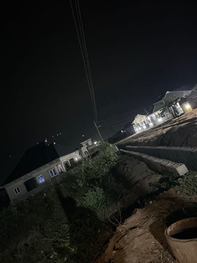 An aerial perspective captures a stark contrast between a black sky and a dimly lit urban landscape. A tall building with faint lights shining through its windows stands out against the darkness. A wall with vegetation sits to the right, and a pipe is placed along the wall's upper edge. Several power lines cut across the black sky above.
