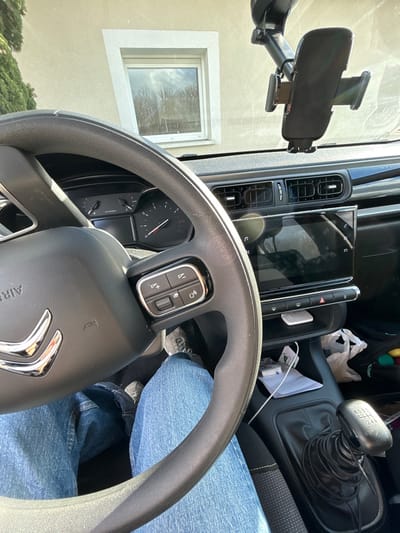 The image displays the interior of a stationary vehicle in Veauchette, France, viewed from the driver's perspective under bright daylight. A person's lower body, wearing blue denim jeans and gray sneakers, occupies the driver's seat. A black multi-function steering wheel with a silver Citroën emblem is prominent in the foreground. Behind the steering wheel, a vehicle instrument cluster with gauges is partially visible. Centrally located on the dashboard is a large, glossy infotainment screen. A black mobile phone mount is affixed to the windshield above the dashboard. To the right and lower in the frame, a six-speed manual gear stick is visible. A storage compartment below the infotainment screen contains a white power bank and a white cable. Through the vehicle's windshield, an exterior light-colored building wall with a white-framed window is visible, reflecting trees and the sky.