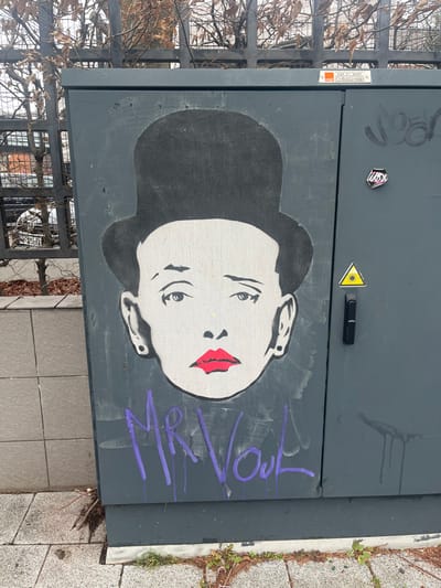 A utility box displays street art. A grayscale portrait of a woman with red lips dominates the surface. The woman has dark hair and pale skin with light shading to show her facial features. A yellow triangle sticker sits above the portrait. Purple graffiti spelling "Mr. Voul" is scrawled beside the face. The box is dark gray. A metal fence and a stone walkway surround the box.