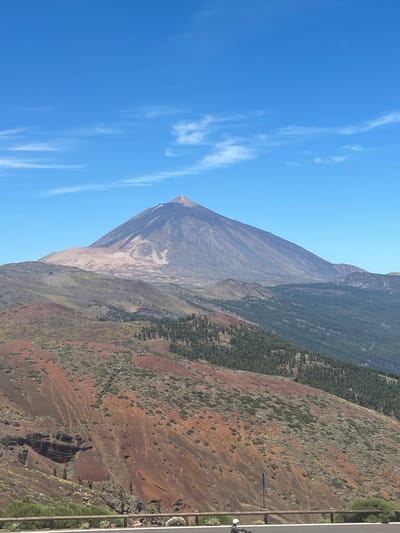 The image shows a landscape dominated by a large, conical volcano under a clear blue sky with scattered white clouds. The volcano's summit is partially obscured by light-colored dust or ash, and its slopes display varying shades of gray and brown. In the foreground, the terrain is characterized by undulating hills covered in red, brown, and green vegetation. Patches of dark green trees are visible on some slopes. A wooden fence runs horizontally across the bottom of the frame, separating the natural landscape from what appears to be a road. A person wearing a white helmet is seated on a dark vehicle on the road. The scene is likely in the vicinity of Teide National Park, which is located on the island of Tenerife in the Canary Islands, and is a prominent natural landmark. While La Orotava is a municipality on Tenerife, the image itself depicts a natural volcanic landscape rather than an urban environment within La Orotava.