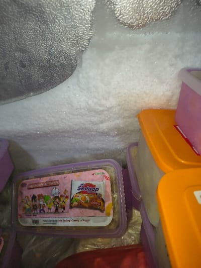 The scene depicts the interior of a freezer compartment within a location in Tarqumiyah, Palestinian Territory. The upper and left portions of the image show a frosted, textured metallic surface. Significant white, crystalline frost accumulation is visible on these surfaces and across the back of the compartment. In the lower half of the image, multiple plastic food storage containers are present. A rectangular container with a translucent purple lid is prominent in the lower-left. Inside this container, a pink-colored item featuring animated characters on the left and a package of "Mie Sedaap Goreng" instant noodles on the right is visible, with accompanying text indicating "Paket komplit Mie Sedaap Goreng isi 4 pcs". To the right of this container, several other food containers are visible, including ones with orange lids stacked atop containers with translucent purple lids. A red object is partially visible at the bottom edge of the frame. No individuals, actions, or direct interactions are depicted.