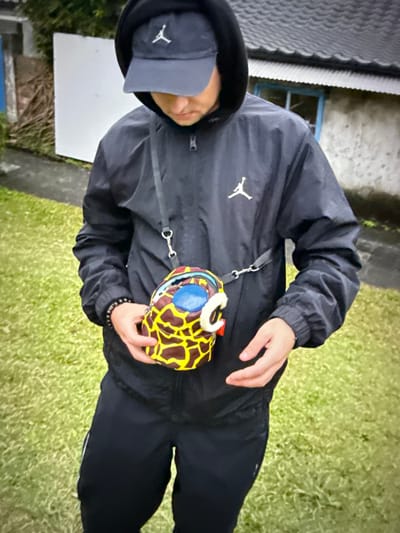 A person wearing a black hooded jacket and a black baseball cap stands outdoors, holding a small, multi-colored bag with a giraffe-like pattern. The person is wearing black pants and has a beaded bracelet on their left wrist. The background consists of a grassy area and a building with a tiled roof and a blue-framed window. The scene appears to be in an urban or suburban environment. No specific location in Taipei, Taiwan, can be identified from the image alone.