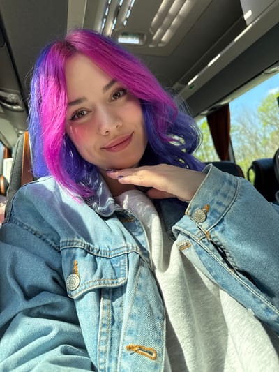 A young woman with striking purple and pink ombre hair is the central focus of this selfie, taken from a slightly high angle and rotated to appear on her side. She looks directly at the camera with a gentle smile. Her makeup includes defined eyebrows, winged eyeliner, and what appears to be pink blush on her cheeks. She is wearing a light grey hooded sweatshirt under a light blue denim jacket. Her right hand is visible near her face, with painted fingernails. The jacket has silver-colored buttons, and one button on the sleeve cuff clearly displays the brand "PULL&BEAR".

The setting appears to be the interior of a vehicle, likely a train or bus, given the long, narrow windows and overhead lighting visible in the upper left corner of the image. Through the windows, bright daylight and green foliage suggest it is daytime, possibly a sunny day. Shadows are cast by the light, indicating a strong light source. The interior of the vehicle is relatively clean and modern, with grey and dark surfaces. The woman appears relaxed, possibly traveling or commuting. No specific activity beyond posing for the camera is evident. The overall mood is casual and bright.