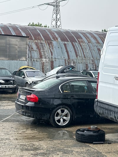 The image shows a collection of cars parked next to a large, weathered metal silo. A white van is parked on top of a black car, its underside visible. The cars appear to be in an outdoor lot, with the ground wet and reflective, suggesting recent rain. Several cars are visible, including a black sedan with its rear left wheel removed and a spare tire resting nearby. The license plate on this car reads "CZ2939BK". Another car, a lighter color, is parked in front of the black sedan, and a portion of its yellow roof is visible. In the background, a large metal silo with rusted corrugated panels dominates the scene. A power line tower and cables are also visible in the distance, along with some greenery. The sky is overcast, indicating a cloudy day. The overall impression is of a car storage or repair yard, possibly in a rural or industrial area. The image was taken in Kazanlak, Bulgaria.