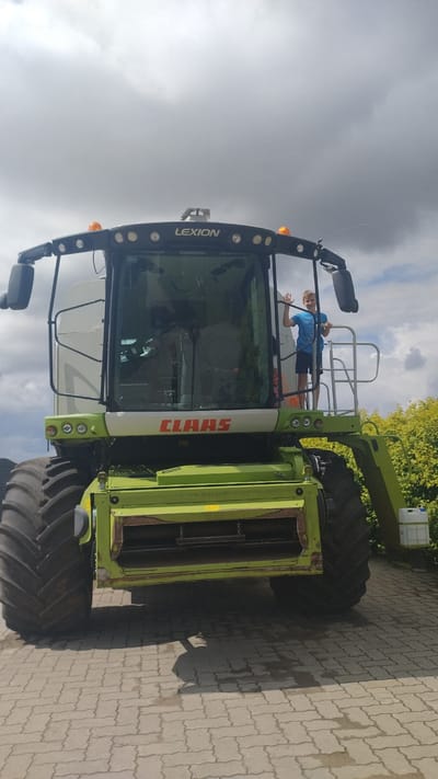 A young boy wearing a blue t-shirt and dark shorts stands on the platform of a large, green Claas Lexion combine harvester. He is waving at the camera. The combine harvester has large, black tires and a prominent Claas logo in orange lettering. The machine is parked on a paved surface, possibly a driveway or farmyard, with a line of green bushes or trees in the background. The sky is overcast with dramatic clouds. The scene appears to be in Kalnrozes, Latvia.