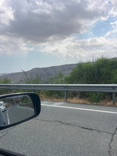 The image depicts a roadside scene, likely in or near Monagri, Cyprus, on a cloudy day. In the foreground, the asphalt road shows visible cracks. A metallic guardrail runs along the edge of the road, separating it from the embankment. To the right of the guardrail, dry grasses and green vegetation are present. In the background, a barren, terraced hillside is visible under a sky filled with substantial gray clouds. The reflection in the car's side mirror shows the road continuing with other vehicles, including a white van.