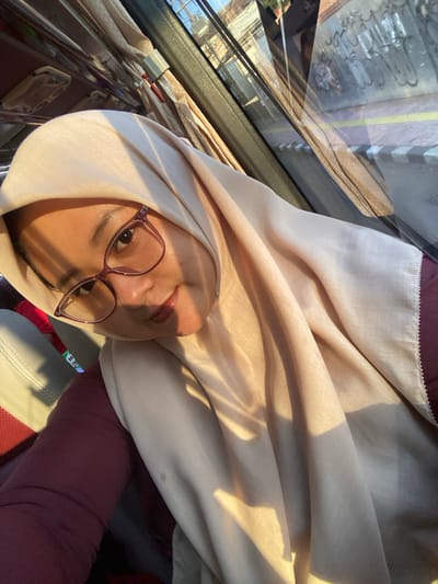 A young adult woman is centered in a selfie taken inside what appears to be a public transport vehicle in Batu, Indonesia. She wears a light beige or cream hijab, plum-framed glasses, and a plum-colored long-sleeved top. Her skin tone is warm, and she has a subtle, tinted lip color. Strong, golden sunlight illuminates the right side of her face and the front folds of her hijab, casting defined shadows. She looks directly at the camera with a gentle, slight smile, her head tilted slightly. To her left, part of a light-colored vehicle seat with red trim is visible. The background, viewed through and reflected in the vehicle window, reveals an urban environment. Visible details include a sidewalk or road edge with a yellow line and a black-and-white striped curb, and a wall with painted patterns or graffiti. Overhead within the vehicle, a light-colored patterned curtain and interior light fixtures are discernible.