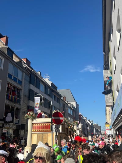 The image shows a street scene in Cologne, Germany, taken from a low angle. The location is near the coordinates 50.926 latitude and 6.958 longitude, which is in the Altstadt (Old Town) district of the city.

The image shows a crowd of people gathered in the street below a line of buildings, possibly during a festive parade. The street is lined with several tall, narrow buildings, all with a gray exterior and multiple windows. The buildings have a slightly aged and worn appearance, with some visible signs of weathering. There are garlands of colorful foliage hanging from the buildings, and some buildings have signs and banners.

At the top of the image, a person dressed in a bright blue jacket can be seen standing on a scaffolding or platform above the street. The person appears to be holding a white object, possibly an umbrella or a large flag. Above the buildings is a bright blue sky with some white clouds visible.

In the street below, there is a crowd of people. It is hard to make out any specific details of the people, as they are tightly packed together and many are wearing colorful costumes and hats. The crowd appears to be lively and energetic.

There are several objects and decorations visible in the scene, including:

* A large, red circle sign with a diagonal line across it, which is a standard "No Entry" sign.
* A large wooden structure in the center of the image, which appears to be a stage or a platform. 
* Several balloons visible in the street and buildings.
* A banner with a "Kleine Knospe" (small bud) label, possibly from a business or restaurant.
* An abundance of colorful decorations and foliage.

The image conveys a sense of festive excitement and energy, with a focus on the brightly colored costumes and decorations of the crowd. The buildings and surroundings suggest a busy, urban location with an active street scene.