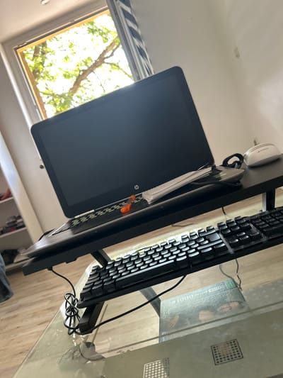 This is a close-up, high-angle, eye-level shot of a laptop, keyboard, and mouse on a black desk, which is positioned on top of a glass table. The laptop, an HP model, is open and its screen is dark. A white face mask and a small orange screwdriver are resting on the laptop's keyboard. To the right of the laptop is a white and gray A4Tech computer mouse. The keyboard is black and appears to be a mechanical keyboard with RGB backlighting, but the lighting is not currently active. Beneath the black desk is a clear glass table. On the glass table, partially obscured by the keyboard and desk, is a puzzle box featuring a depiction of Leonardo da Vinci's "Mona Lisa". Behind the desk, a window is visible, showing bright green trees and sunlight filtering through. The wall to the right is painted a light color and appears to have two small, circular marks. The flooring is a light-colored wood. The overall scene suggests a home office or study area. This image was taken in Schöningen, Germany.