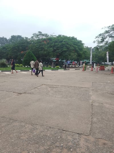 This image depicts an outdoor public space, likely a large plaza or courtyard, within the city of Ota, Nigeria, under an overcast sky. The foreground and midground consist of worn, grey asphalt or concrete paving, exhibiting cracks and exposed aggregate.
In the left midground, a female individual in a dark dress and red shoes walks from left to right. Behind her, three male individuals proceed in the same direction: one in a light patterned shirt and dark pants, another in a plaid shirt carrying a purple bag, and a third in a light patterned shirt and dark pants.
Further in the midground, to the right, several orange traffic cones are positioned on the paved surface near a rectangular barrier. Beyond these, a landscaped area features green grass, low bushes, a conical evergreen, and a spiky plant, demarcated by a low white curb. Within this area and slightly behind it, multiple indistinct figures are visible, including one holding a red umbrella and another in a blue shirt.
The background is dominated by dense green foliage of large trees, some displaying clusters of red flowers. Tall, white, pillar-like structures are also present on the right side of the background.