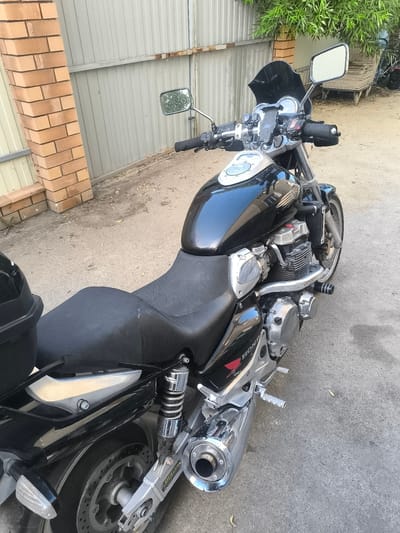 A black Honda motorcycle is parked on a paved surface, likely a driveway or street. The motorcycle is positioned at an angle, facing towards the right of the frame. It has a sleek, modern design with chrome accents and a black windshield. The seat appears to be black, with a small tear visible on the right side. The engine is exposed, showcasing its mechanical components. The motorcycle is adorned with the Honda logo on its fuel tank.
In the background, a corrugated metal fence runs horizontally across the image. To the left of the motorcycle, a brick wall is visible, suggesting a residential or industrial setting. Trees with green foliage can be seen behind the fence on the right side of the image, along with a wooden structure that appears to be a bench or a pallet stacked with wood. The ground around the motorcycle is a mix of dirt and fallen leaves. The scene is illuminated by natural daylight, casting soft shadows. The location is identified as Kopeysk, Russia. There are no people visible in the image.