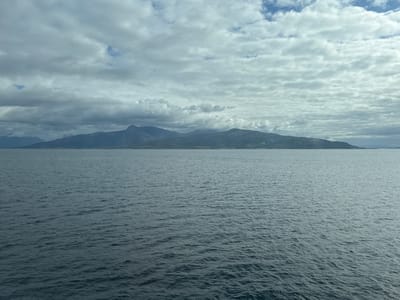 The image depicts a vast expanse of calm, dark blue water in the foreground, reflecting the overcast sky. In the distance, a long, low-lying island or peninsula with rolling hills and a mountain range stretches across the horizon. The mountains are covered in shadow, with some peaks emerging from low-hanging clouds. The sky above is a mosaic of grey and white clouds, with occasional patches of lighter blue peeking through. The overall scene is one of natural, serene beauty, characteristic of the Norwegian landscape. This view is likely from the waters surrounding the town of Lodingen, Norway.