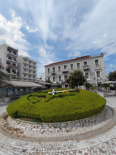 The image depicts a well-maintained outdoor public space in Patras, Greece, featuring a large, artfully shaped green hedge, likely a flowerbed or a topiary, with yellow flowers interspersed. The hedge is circular in shape and is surrounded by a paved area made of small, multi-colored stones laid in a radial pattern. Behind the hedge, a light-gray building with numerous windows and balconies stands tall. Further back, another modern building with white balconies is visible. The sky is a bright blue with scattered white clouds, suggesting fair weather. There is no specific activity or event taking place; the scene appears to be a snapshot of an urban landscape. Text is visible on the buildings, including signs with Greek lettering. One sign on the light-gray building reads "ΑΓΙΟΥ ΓΕΩΡΓΙΟΥ" and another appears to be for a business named "ΠΙΤΣΑ". The overall impression is one of a clean, well-kept urban environment with architectural elements and greenery. The time of day appears to be daytime, given the bright lighting and clear sky.