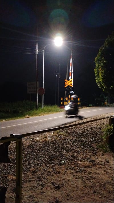 This is a night-time, eye-level shot of a road crossing railroad tracks in Tulungrejo, Indonesia. A street lamp illuminates the scene, casting bright light and creating lens flare. A train crossing gate with red and white stripes and reflective yellow Xs is lowered, indicating an approaching train or ongoing crossing. A red octagonal stop sign is posted to the left of the road. The road itself is paved with a white line dividing the lanes. A motorcycle is captured in motion on the road, appearing blurred due to its movement. The area to the right of the road is covered in gravel and dirt, with grass and weeds growing through. To the left of the road, there is a grassy embankment. A tree with green foliage is visible on the far right of the image. A sign with Indonesian text is mounted on a pole in the background. The sky is dark, with some faint light streaks that could be from light pollution or lens artifacts.