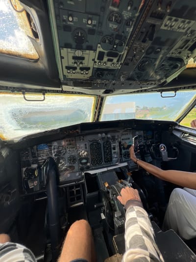 The image shows the interior of an aircraft cockpit, viewed from a slightly elevated and tilted perspective. The cockpit is filled with numerous analog instruments, gauges, switches, and controls, characteristic of older aircraft. The overall impression is one of a functional, though perhaps slightly dated, cockpit environment.

In the foreground, two people are visible. One individual, seen from the chest down, is wearing a black and white checkered long-sleeved shirt and is operating a control lever in the cockpit. Another person, wearing white pants, has their arm reaching towards the control panel, holding a smartphone, possibly taking a picture or video. The cockpit windows are visible, showing a bright, sunny outdoor scene with greenery and buildings, suggesting it is daytime. The windows appear somewhat dirty or weathered, with water or condensation on the left window.

The visible text includes labels for various aircraft systems such as "EMER EXIT LIGHTS", "FASTEN BELTS", "SMOKING", "FUEL PUMPS", "CROSS FEED", "IPS", and "BUS TRANSFER". There is also a visible aircraft identification number "N25545".

The scene appears to be a stationary aircraft, possibly on the ground. The people's interaction with the cockpit suggests they are exploring or documenting the interior. The setting in Blahbatuh, Indonesia, implies a tropical or subtropical climate, which is consistent with the bright daylight and greenery seen through the windows. There are no obvious cues about the emotional state of the individuals, but their engagement with the cockpit suggests curiosity or interest.