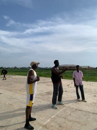 In Gwafan, Nigeria, a basketball court scene unfolds under a partly cloudy sky. Three men stand on the cracked concrete court. The man in the foreground, wearing a white and yellow basketball jersey, a tan baseball cap, and black socks with slides, faces right, his body angled as if in conversation. To his right, a man in a black sleeveless shirt and grey pants gestures with his right hand, pointing towards something beyond the frame, his gaze directed forward. Beside him, a third man in a pink button-down shirt and dark jeans stands facing forward, a slight smile on his face. In the background, a lone figure in a dark jacket and shorts walks away from the camera. Beyond the court, a field of green grass leads to a row of long, brick buildings with light-colored roofs, suggesting an institutional or residential area.