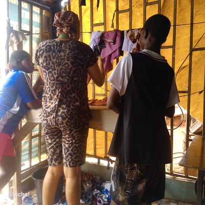 Three people are inside what appears to be a tailor's shop in Nsugpe, Nigeria. The person in the center is wearing a matching leopard print top and shorts and has a scarf wrapped around their head. They stand at a table, appearing to work with fabric. To their right, a person wearing a white and black outfit stands looking towards the table. To the left, a third person in a blue top is leaning on the table, observing. Sewing supplies, materials, and a bucket are visible below the table. A yellow fabric backdrop partially blocks the outside view. The shop appears to be contained behind metal bars, and another table is seen to the right, with a sewing machine beneath it.