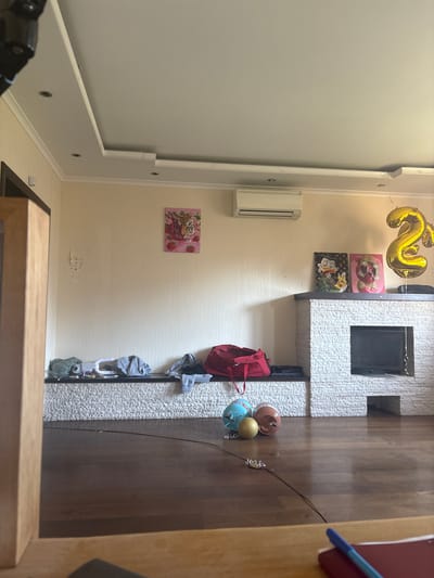 The image depicts a dimly lit living room with a wooden floor and light-colored walls. In the center of the room, a fireplace made of white stone is visible, with a black insert. Above the fireplace, two framed pictures are hung, one of Scrooge McDuck holding a Bitcoin symbol and another of a character resembling a pig wearing sunglasses. A large, gold balloon in the shape of the number "2" is floating to the right of the fireplace.
In front of the fireplace, on a white, textured surface, there are several items including a red duffel bag and some folded clothing. On the wooden floor, in front of this surface, are three round objects: a blue and turquoise sphere, a brown sphere, and a gold sphere. A thin black wire extends across the floor from the left side of the frame, appearing to be attached to the gold sphere with a small white object.
To the left of the fireplace, a white air conditioning unit is mounted on the wall. Above the white textured surface, a framed picture of a tiger with strawberries is hung on the wall. A wooden furniture piece is partially visible on the far left of the image, and a notebook with a blue pen is in the foreground on a wooden surface. The overall impression is of a domestic setting, possibly prepared for or recently having had a celebration, given the balloon and decorative items. The location is specified as Orekhovka, Russia.