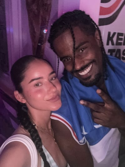 In Miami Beach, Florida, a Black man with braided hair and a man with brown hair and braided hair are captured in a selfie. The man with dark hair is wearing a blue jersey with white and red trim. He is smiling and making a peace sign with his right hand. The woman in the image has her hair in a braid and is wearing a white top. Her face is slightly flushed, and she is looking at the camera. The background is blurred, with hints of purple lighting and a wall with text that appears to say "KEN" and "TAS". The overall impression is a casual and friendly atmosphere.